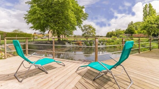 Een houten terras met twee turquoise ligstoelen gericht op een vijver, omgeven door groen en een heldere lucht.