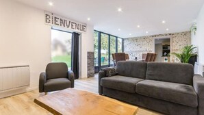 Een moderne woonkamer met een grijze bank, een houten salontafel en een zwarte fauteuil. Op de muur staat een bord met daarop 'BIENVENUE'.