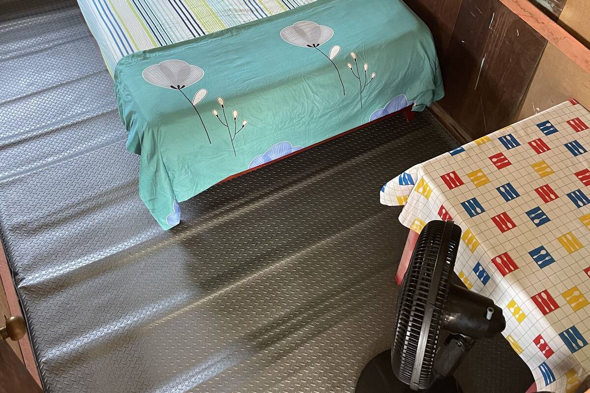 Ein Einzelbett mit gestreifter Bettwäsche, einem grünen Kopfkissen und einem Kopfteil aus Metall. Ein Ventilator steht auf dem Boden neben dem Bett.