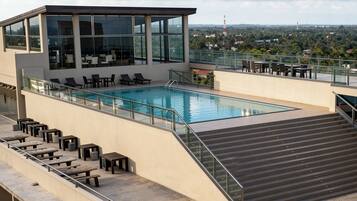 Piscina en la azotea con sillones reclinables y mesas, rodeada por una barandilla de cristal y vistas a la ciudad.