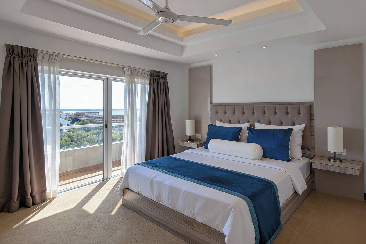 A hotel room with a large bed, a ceiling fan, a nightstand with a lamp, and a view of the city through the window.