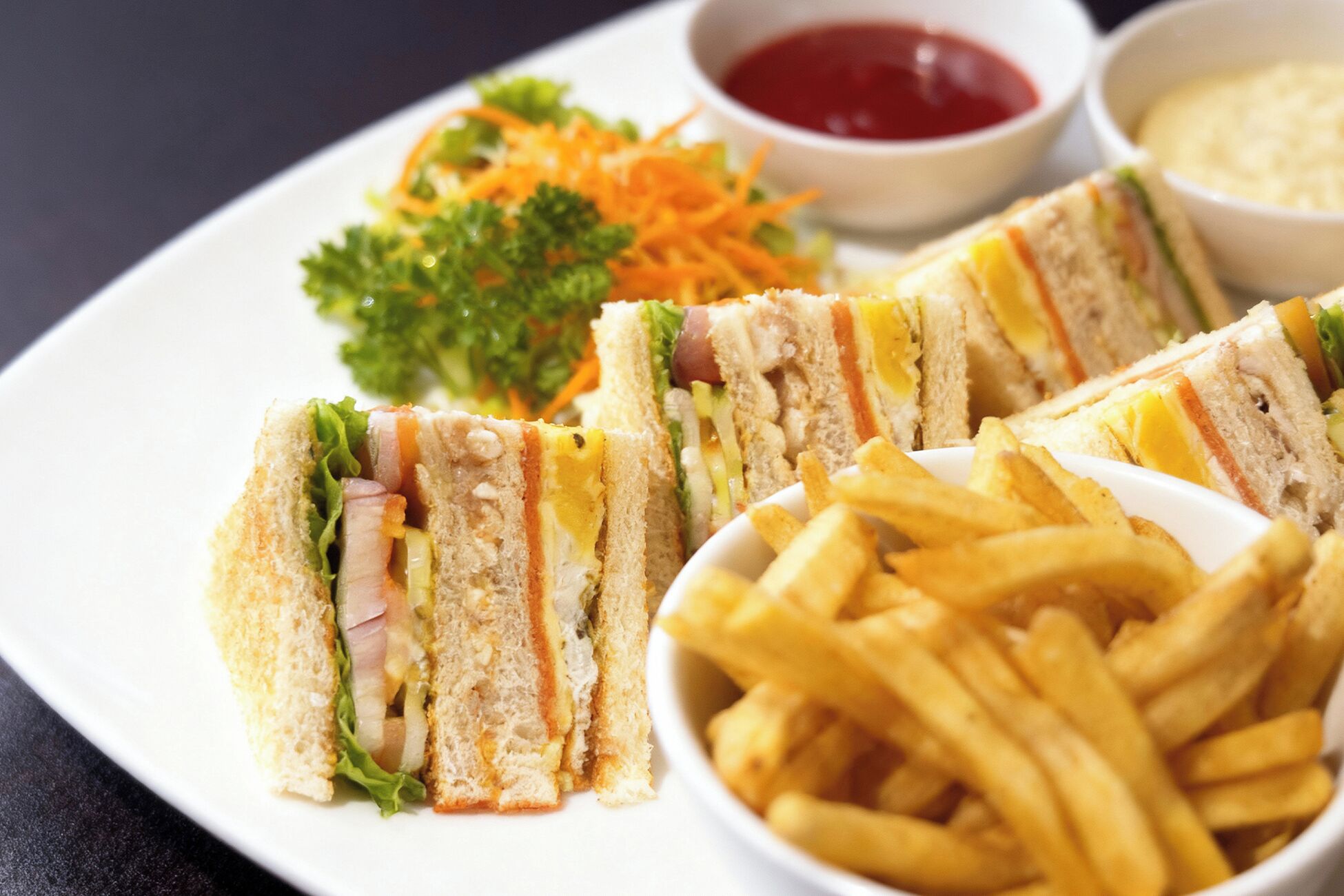 A plate with a sandwich cut into four pieces, a side of French fries, and two small bowls containing sauces.