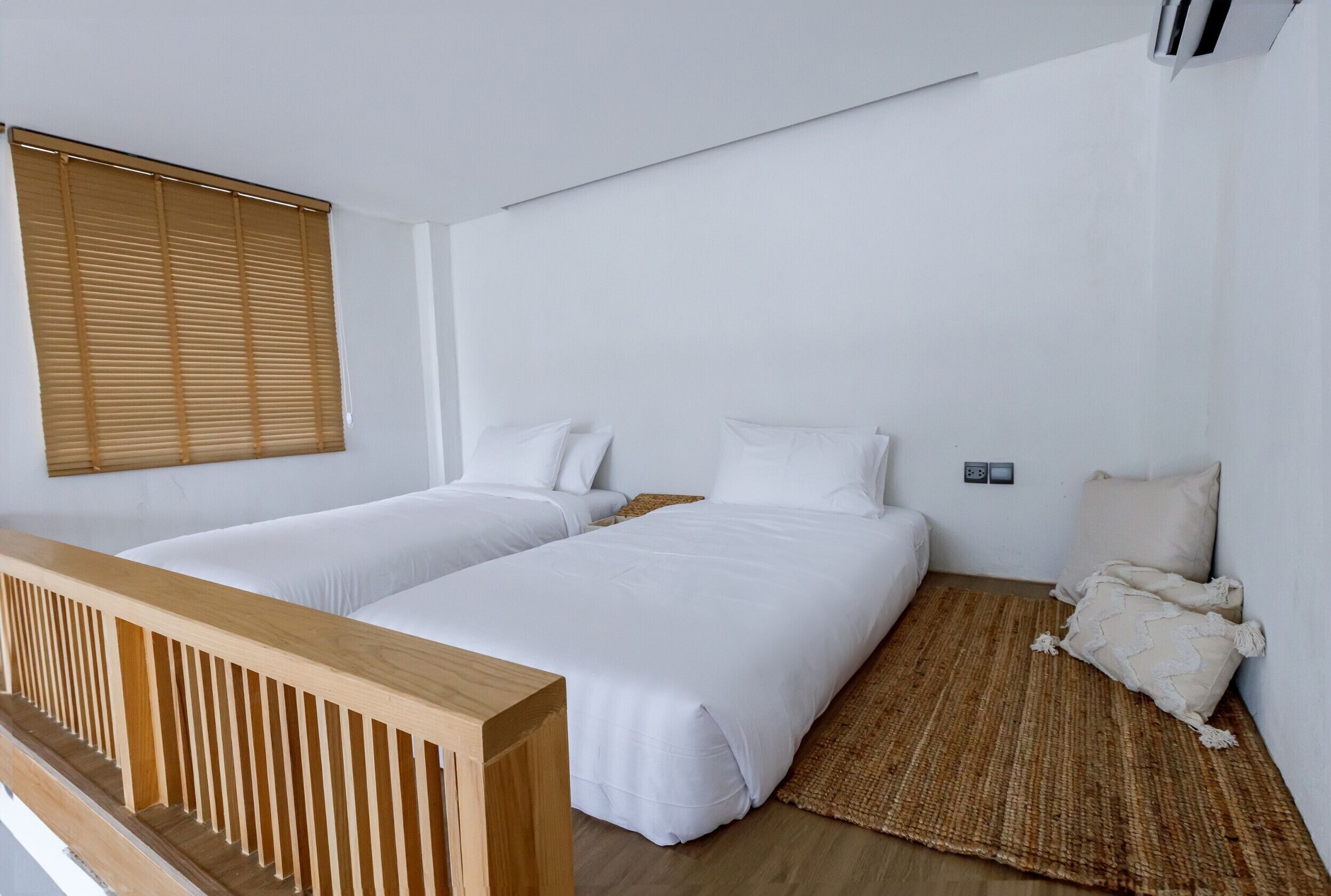 Ein Zimmer mit zwei Einzelbetten, einem Holzkopfteil, einem gewebten Teppich und einem Fenster mit Jalousien.