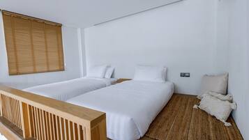 Ein Zimmer mit zwei Einzelbetten, einem Holzkopfteil, einem gewebten Teppich und einem Fenster mit Jalousien.