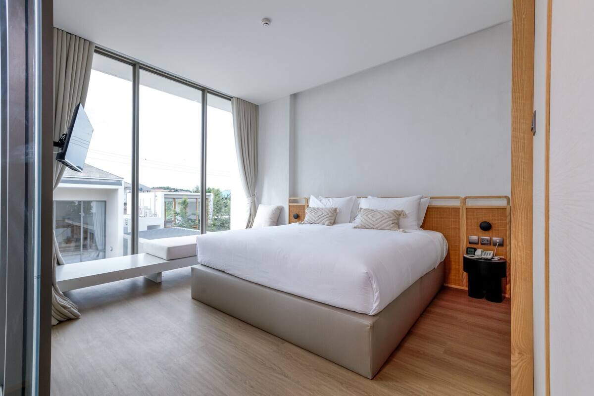 A modern hotel room with a large bed, a small bedside table, a nightstand, and a large window with curtains.