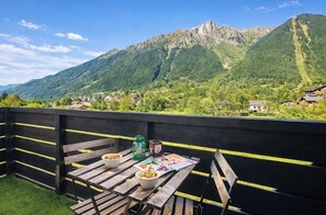 A wooden table with bowls of food, a green water bottle, and a magazine on a balcony with a mountain view. - Cosy studio with view of Brévent (Chamonix-Mont-Blanc)