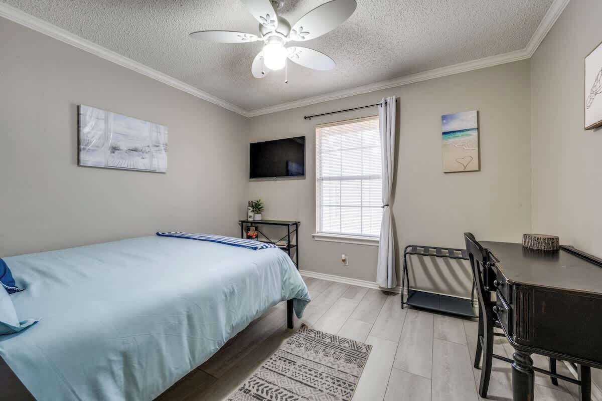 Une chambre à coucher comprenant un lit, un bureau, une chaise, un ventilateur de plafond, une télévision et une fenêtre avec des rideaux.