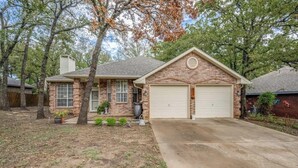 A single-story brick house with a garage, a front yard with trees and plants, and a concrete driveway. - Spacious Brick House 3BR 2BA A+ (Denton)