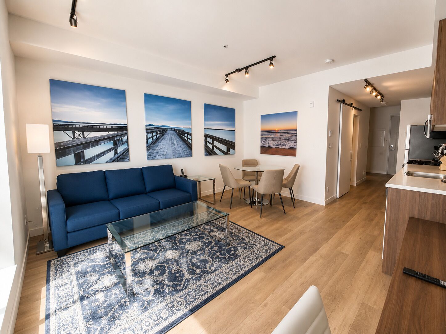 A modern living room with a blue sofa, a glass coffee table, and a patterned rug. There are three framed pictures on the wall, a dining area with a table and chairs, and a kitchen area with a microwave and stove.