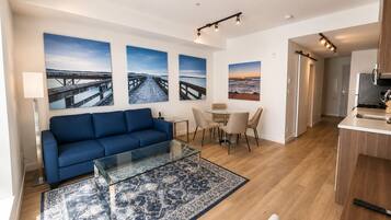 A modern living room with a blue sofa, a glass coffee table, and a patterned rug. There are three framed pictures on the wall, a dining area with a table and chairs, and a kitchen area with a microwave and stove.