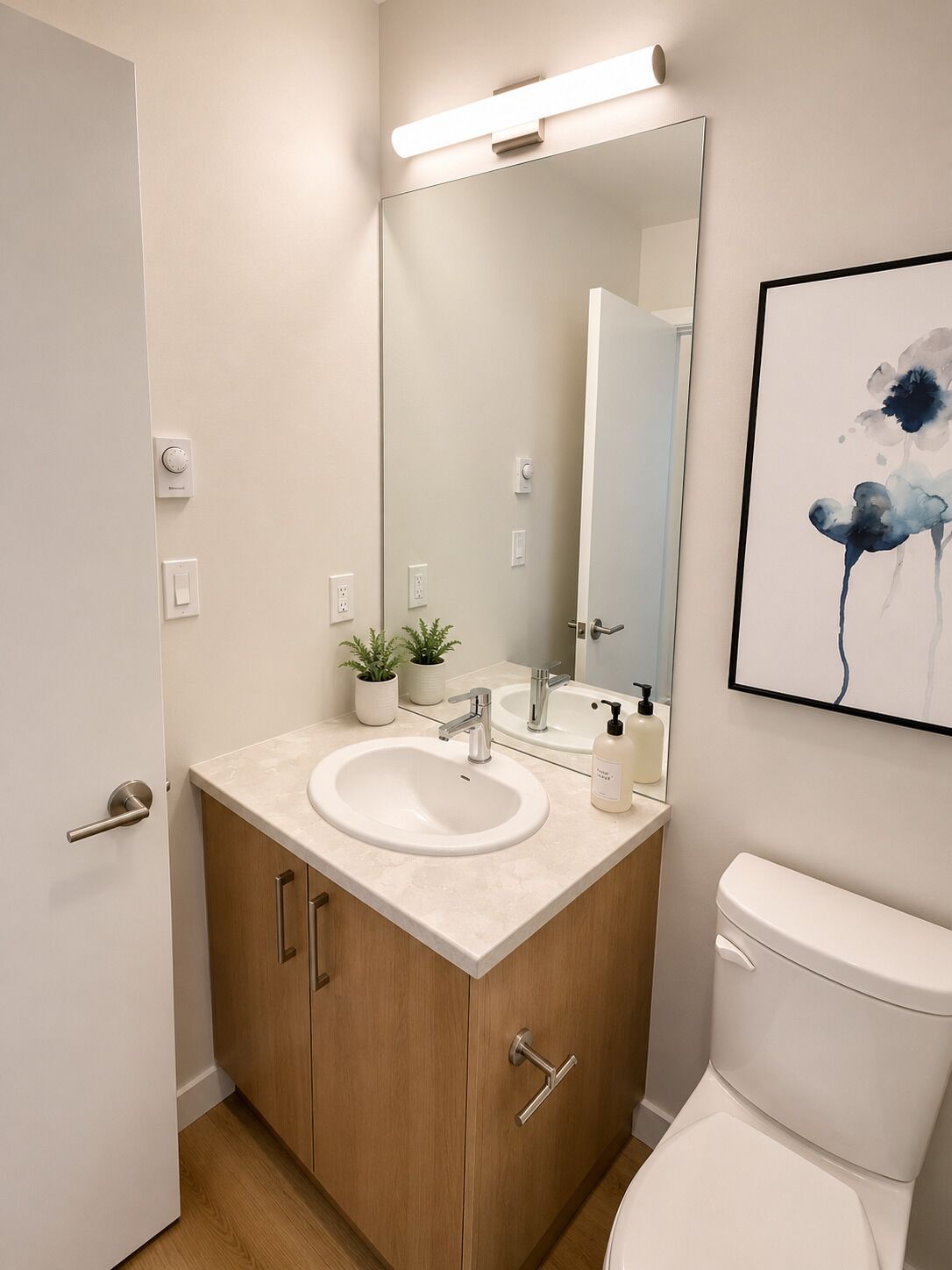Un baño moderno con un lavamanos blanco, armarios de madera, un inodoro, un espejo grande y una obra de arte enmarcada en la pared.