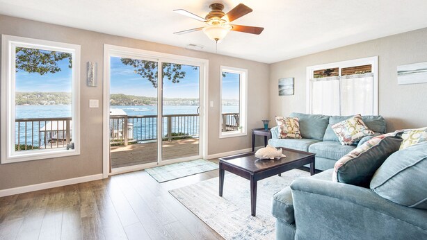 A living room with a view of the water, a ceiling fan, and a large window. - Two Homes, Large Deck, Firepit, Central Location (Sunrise Beach)