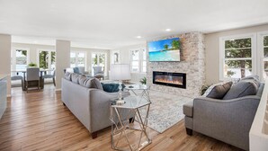 A spacious living room with a fireplace, a large TV, and a dining area with a table and chairs. - Two Homes, Large Deck, Firepit, Central Location (Sunrise Beach)