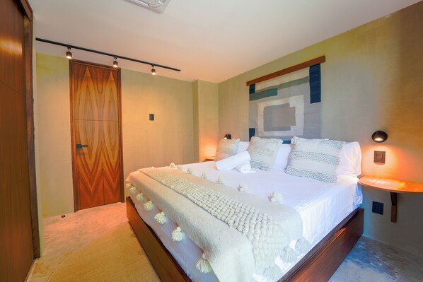 A neatly made bed with a white comforter and pillows, a wooden headboard, and a wall-mounted lamp. - Maramara Centro (Sayulita)