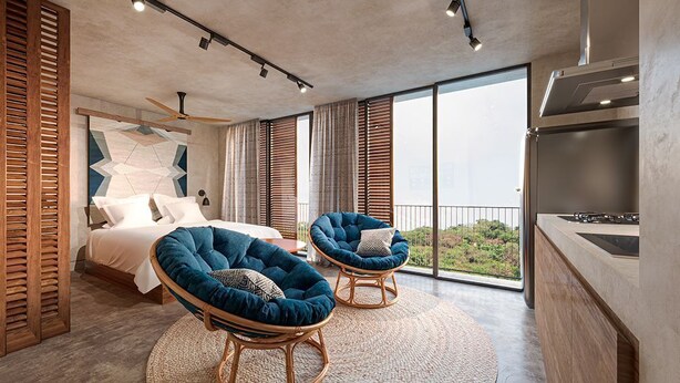 Une chambre moderne avec un grand lit, deux fauteuils bleus, un balcon avec vue et un coin cuisine.