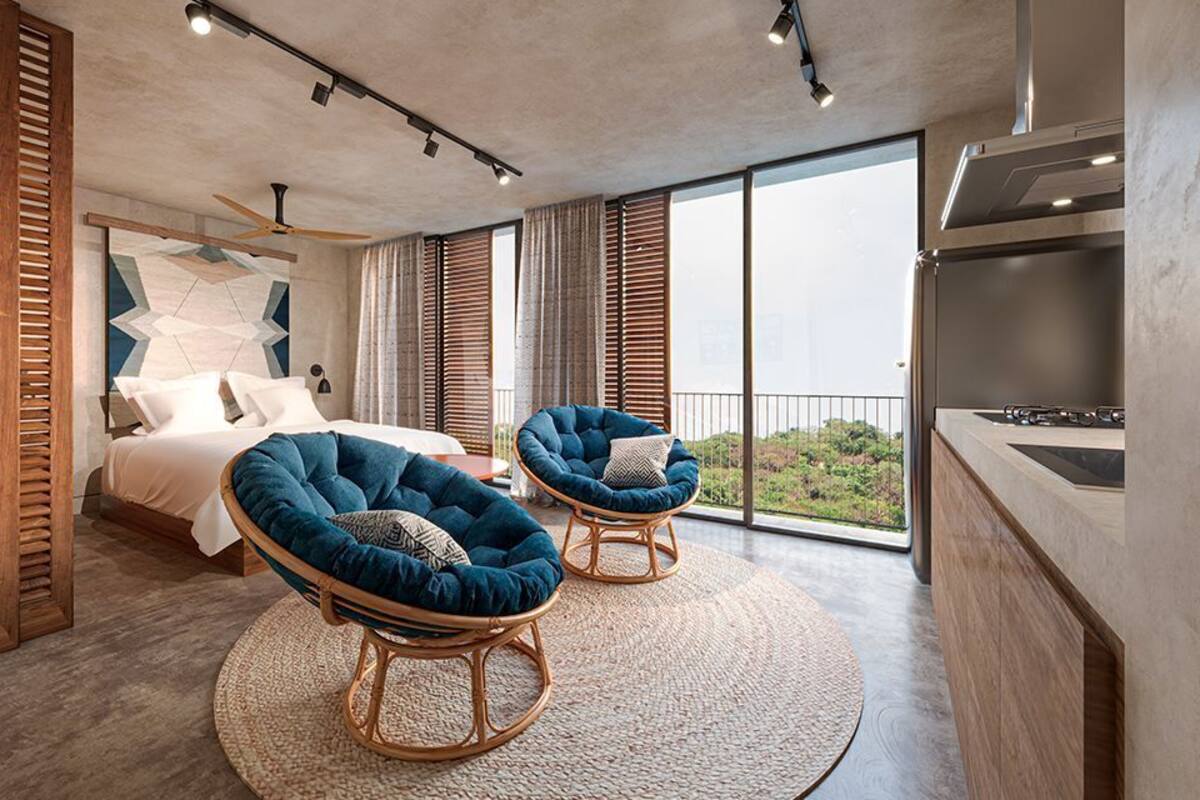 Une chambre moderne avec un grand lit, deux fauteuils bleus, un balcon avec vue et un coin cuisine.