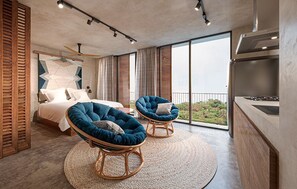 A modern bedroom with a large bed, two blue armchairs, a balcony with a view, and a kitchen area. -  Maramara Centro (Sayulita)