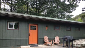Uma casa com fachada verde, porta vermelha, duas cadeiras Adirondack e uma churrasqueira.