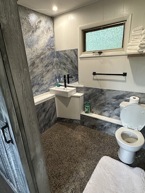 Une salle de bain avec un mur en marbre, un lavabo blanc, des toilettes et une fenêtre située au-dessus des toilettes.