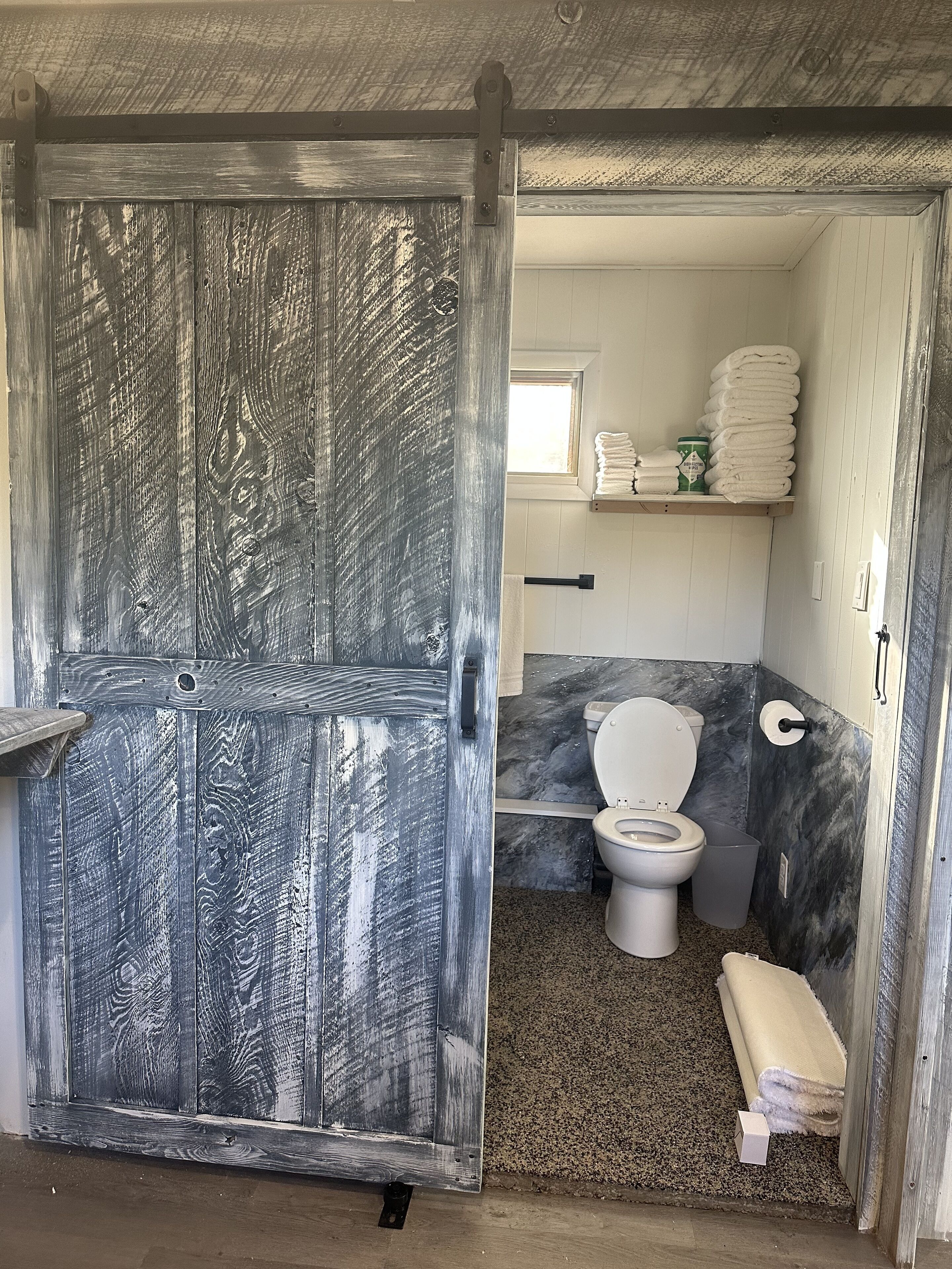 Une salle de bain comprenant des toilettes, un lavabo et un espace douche. On y trouve un porte-serviettes avec des serviettes et un récipient vert.
