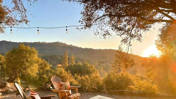 A patio with wooden chairs and a table, overlooking a hilly landscape with trees and a sunset. - Sierra National Forest & Yosemite Estate + Orchard Retreat (Oakhurst)