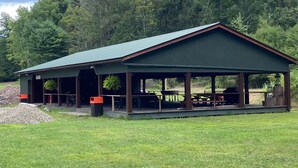 Een overdekte buitenpaviljoen met picknicktafels en bankjes, omringd door gras en bomen.