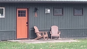 Een gebouw met een rode deur, twee Adirondack-stoelen en een kleine tafel buiten.