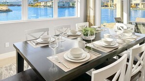 A dining table set for six with white chairs, positioned by a large window overlooking a body of water and buildings. - Casa Marina -15 min to Coachella (Indio)