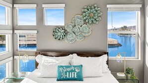 A bedroom with a large bed, multiple windows, a view of a body of water, and decorative wall art. - Casa Marina -15 min to Coachella (Indio)