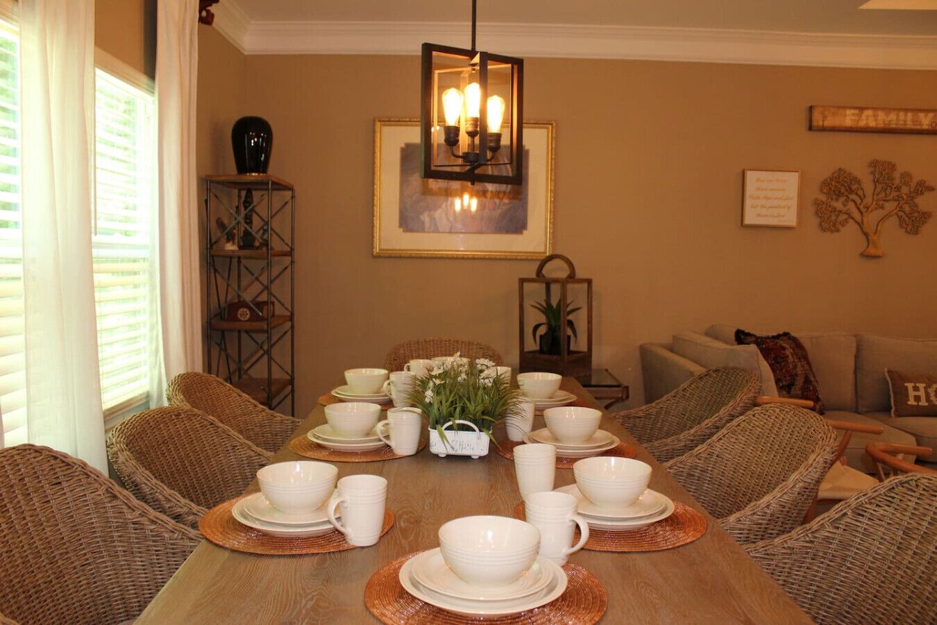 A dining room with a wooden table set for a meal, surrounded by wicker chairs, and a chandelier above.