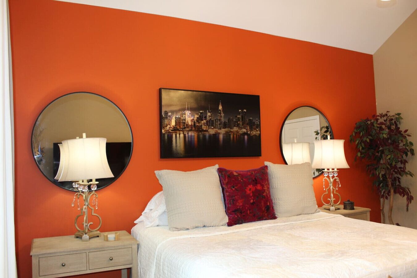 A bedroom with a bed, two bedside tables, a nightstand, a mirror, a painting, and a potted plant.