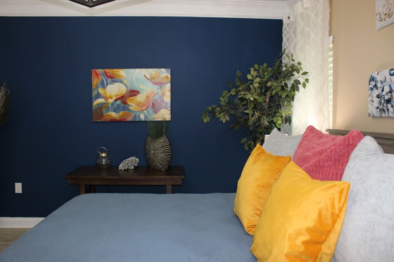 Een slaapkamer met een blauwe spreidek, een decoratief schilderij, een kleine tafel met een vaas en een theepot, en een potplant.