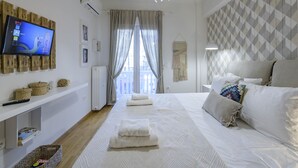 Una habitación de hotel moderna con una cama grande, un televisor de pantalla plana montado en un panel de madera, un aire acondicionado de pared y vistas a la ciudad a través de cortinas translúcidas.