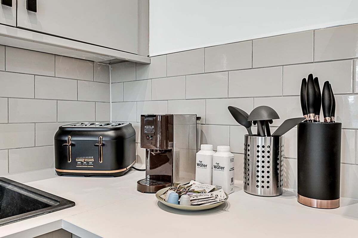 A kitchen with a black toaster, a coffee maker, two bottles of water, a metal utensil holder, and a plate with newspapers.