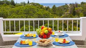 Une table de petit-déjeuner avec des fruits, des croissants et du jus, offrant une vue magnifique sur l’océan et la végétation environnante.