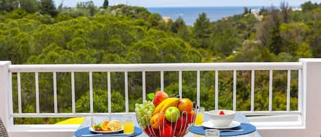 A breakfast table with fruit, croissants, and juice, overlooking a scenic view of the ocean and greenery.