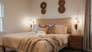 Una cama bien hecha con una manta color beige y almohadas, una mesita de noche con un florero y decoración en la pared.