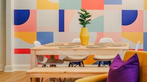 Un área de comedor con una mesa de madera, sillas blancas, un jarrón amarillo con una planta y un cojín morado en un sofá.