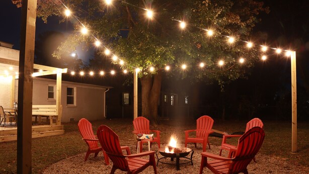 Un patio trasero con un brasero rodeado de sillas Adirondack rojas, luces colgantes y una pérgola de madera.