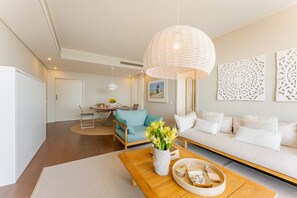Uma sala de estar moderna com uma mesa de centro de madeira, um sofá azul e um sofá branco. Ao fundo, há uma área de jantar com uma mesa e cadeiras.