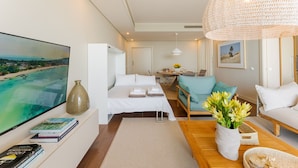 A modern hotel room with a large flat-screen TV, a bed with white linens, a wooden coffee table, a sofa, and a large woven light fixture. - FLH Seaside Luxury at Madeira Palace Residence (Funchal)