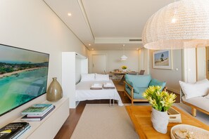 Quarto de hotel moderno com uma televisão de tela plana grande, uma cama com roupa branca, uma mesa de centro de madeira, um sofá e um luminário tecido grande.
