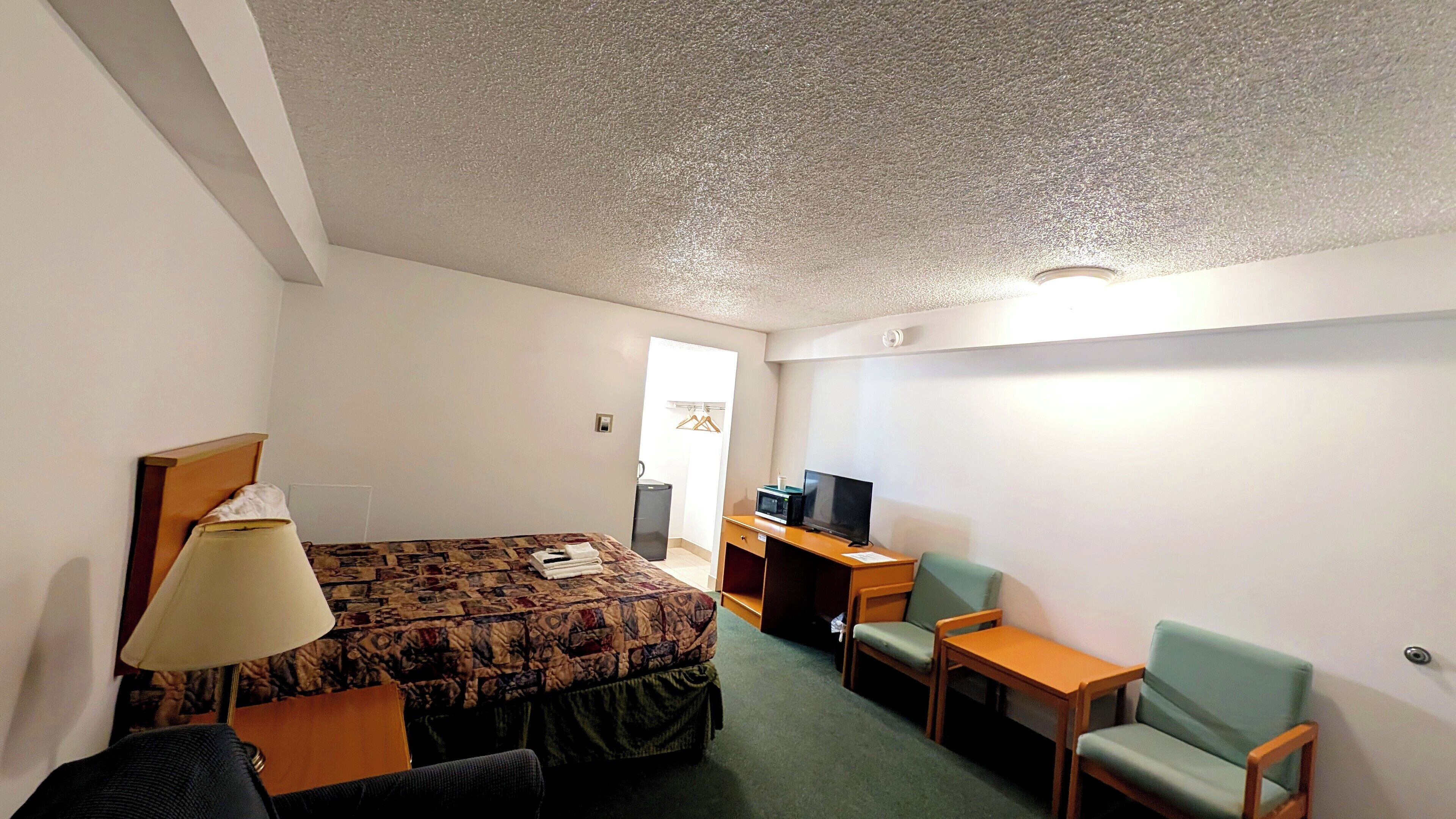 A hotel room with a bed, a desk with a computer, a TV, two chairs, and a lamp.