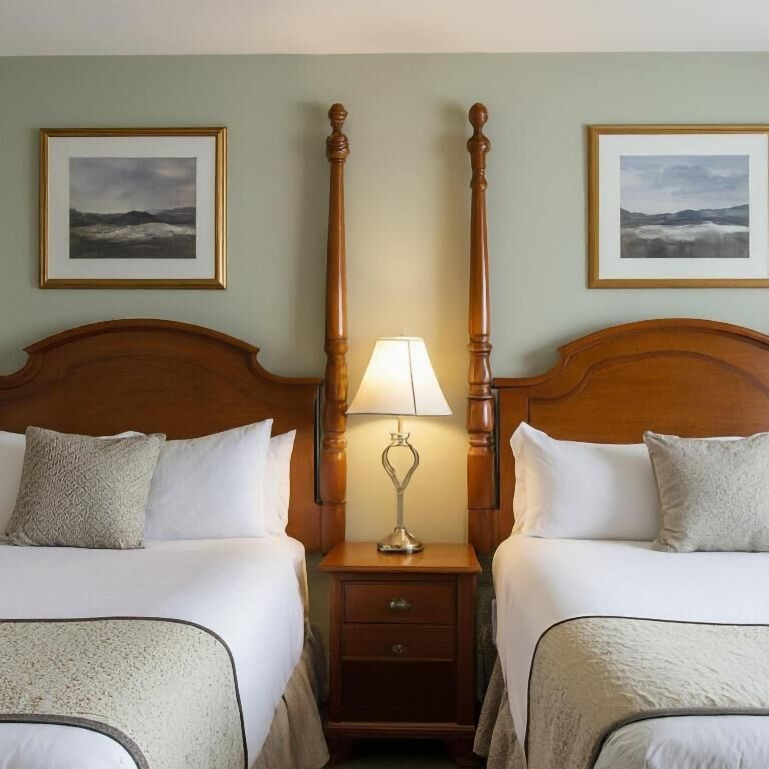 A hotel room with two beds, a nightstand with a lamp, and two framed pictures on the wall.
