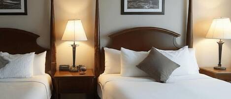 A hotel room with two single beds, each with a wooden headboard, white bedding, and a bedside table with a lamp. There are framed pictures on the wall above each bed.