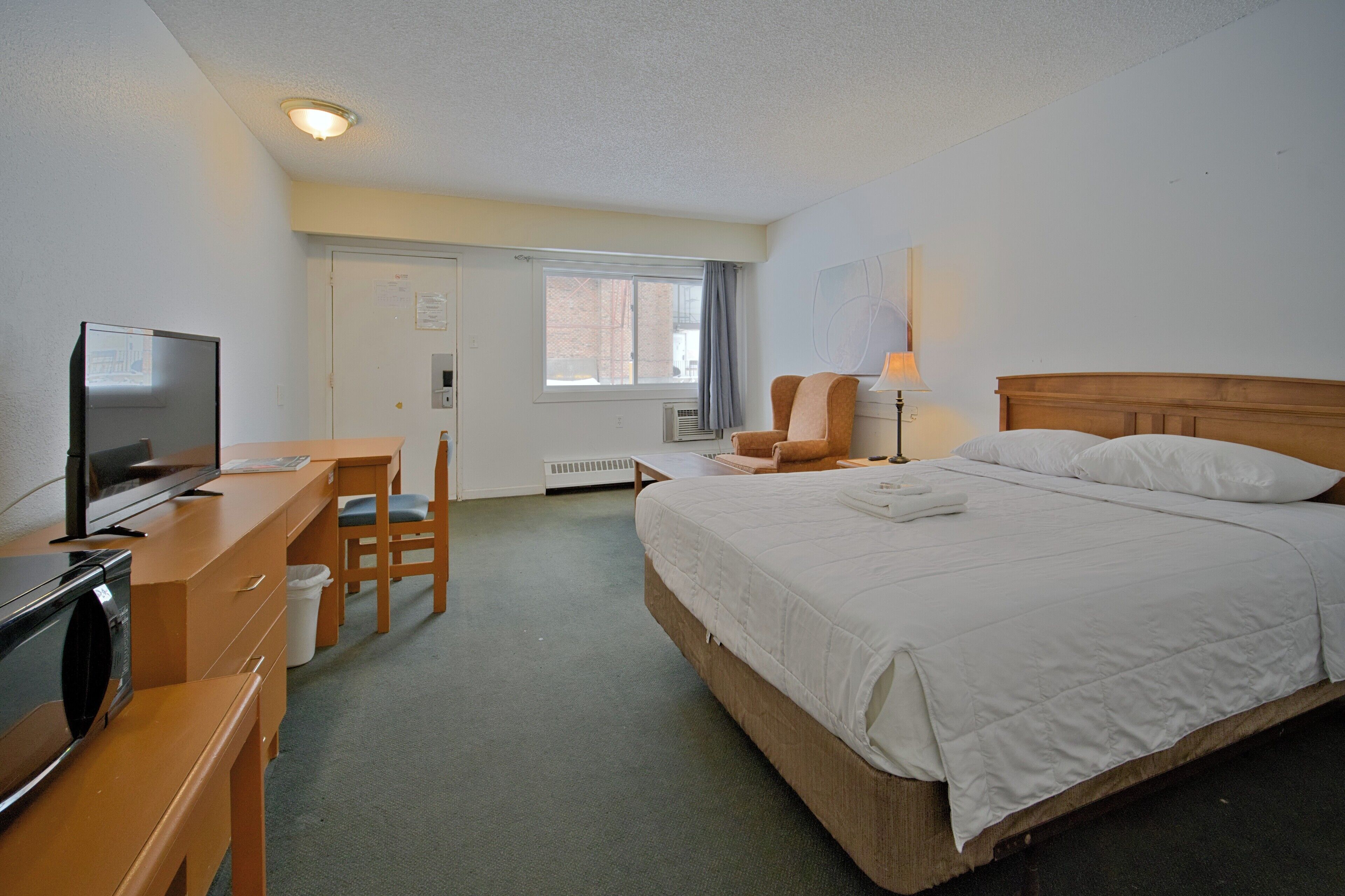 A hotel room with a bed, a desk with a television, a chair, a lamp, and a window.