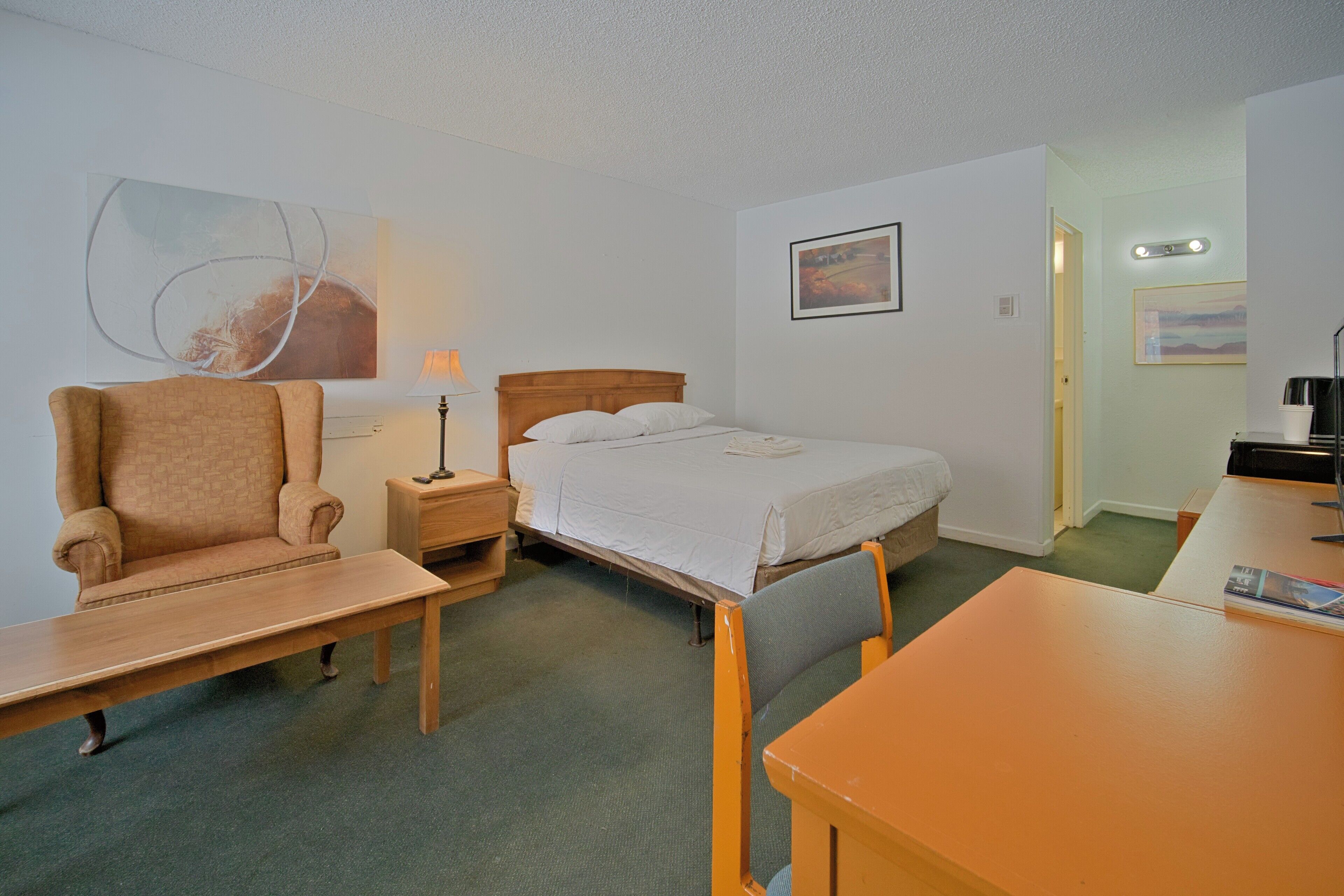 A hotel room with a bed, a chair, a table, a lamp, and artwork on the walls.