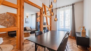 A spacious living area with a large wooden dining table, a sofa, and a coffee table. There are hanging light fixtures and a view of the outside through large windows.