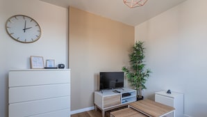 A modern, minimalist living room with a flat-screen TV, a white cabinet, a wooden table, a potted plant, and a wall clock. - L'Escapade Strasbourgeoise - Magnifique petit T2 refait à neuf (Strasbourg)