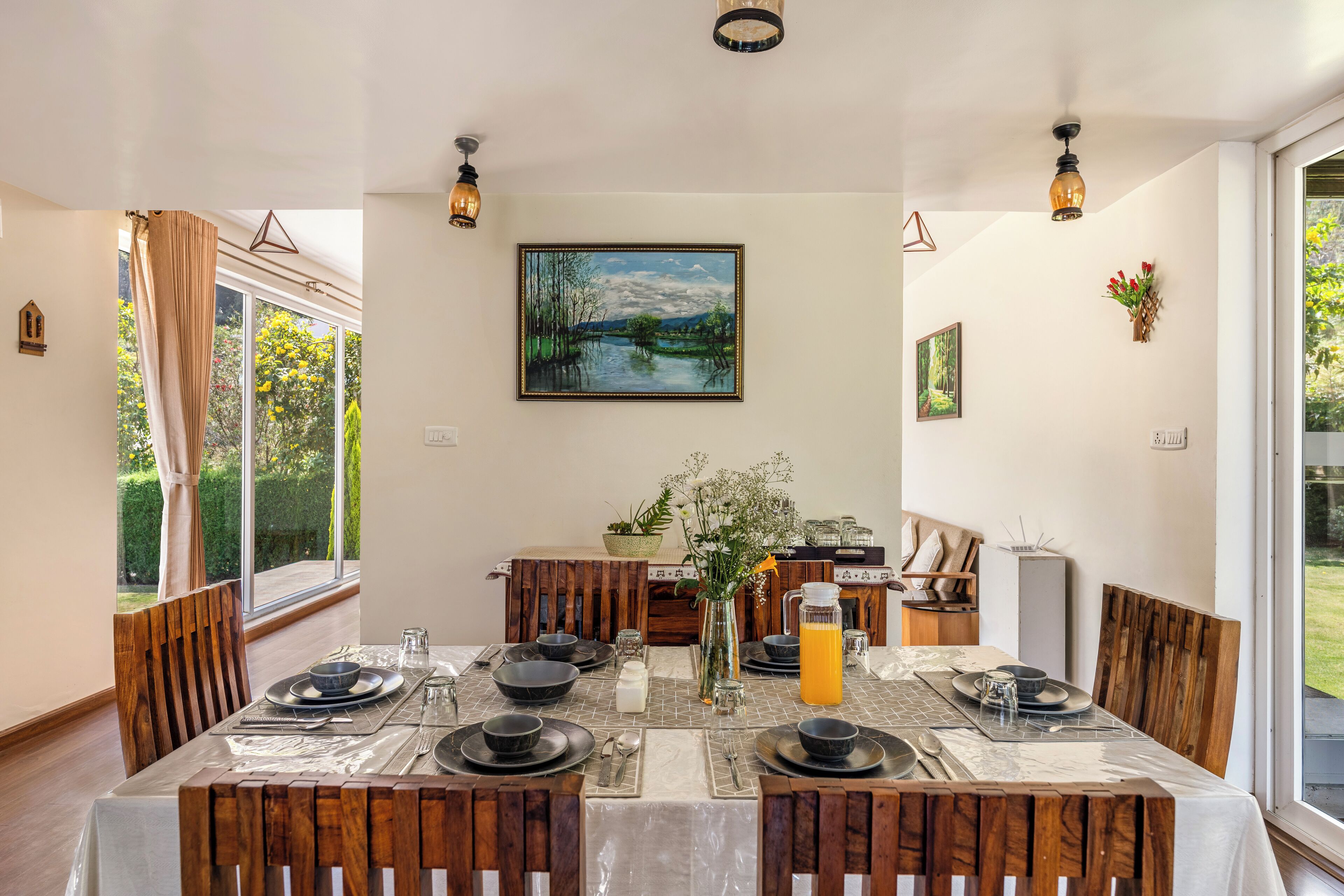 A dining area with a table set for four, surrounded by wooden chairs, a vase with flowers, and a painting on the wall.